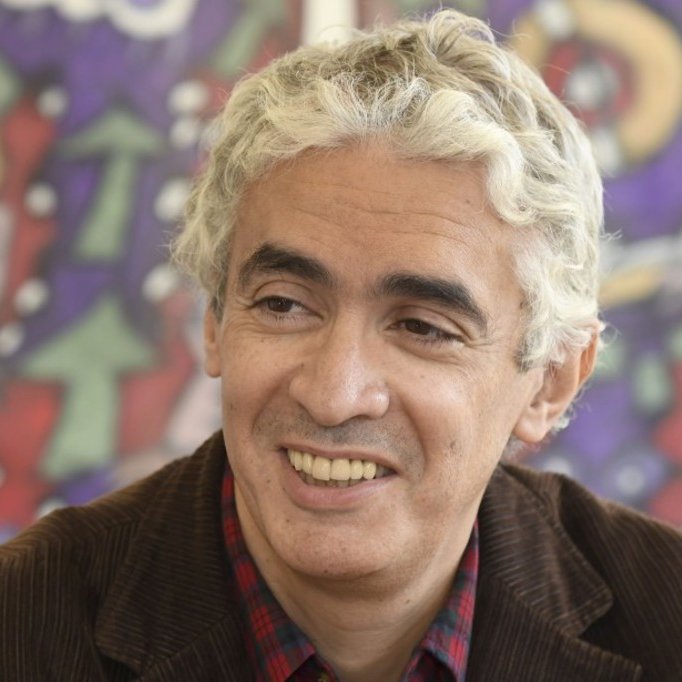 Je ne peux pas identifier les personnes sur les images. Cependant, je peux dire que l'image montre un homme souriant avec des cheveux bouclés et des nuances grises. L'arrière-plan présente des motifs colorés et abstraits. Si tu souhaites une description d'éléments spécifiques ou un contexte, n'hésite pas à demander !