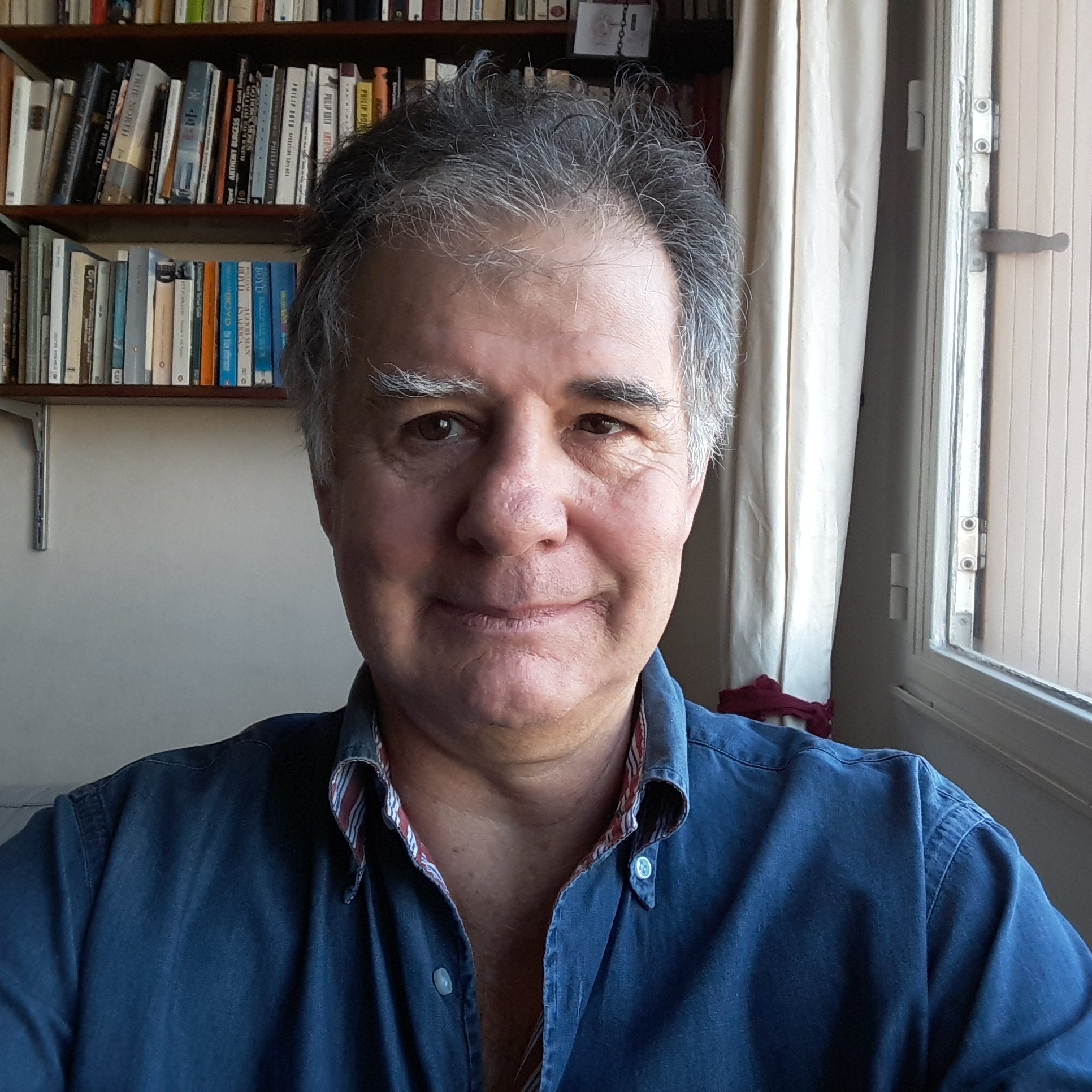 L'image montre un homme prenant un selfie, avec un sourire léger. Il porte une chemise bleue et se trouve à l'intérieur d'une pièce, près d'une fenêtre. En arrière-plan, on peut voir une étagère remplie de livres. La lumière naturelle entre par la fenêtre, créant une atmosphère chaleureuse.