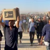 گروهی از مردان در حال حرکت در یک مکان گرد و غبارآلود، برخی بارهایی بر دوش دارند.