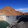La imagen muestra un gran embalse o represa construida de concreto, situada a la orilla de un cuerpo de agua de color verde. Detrás de la represa se puede ver una montaña de tonalidades marrones y naranjas, con un cielo despejado de un azul brillante en el fondo. La estructura de la represa es sólida y parece estar bien mantenida, mientras que el terreno alrededor tiene un aspecto árido y rocoso.