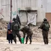Un grupo de soldados está registrando una bolsa cerca de una mujer en un entorno urbano deteriorado.