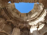 L'image montre l'intérieur d'une structure ancienne, probablement un temple ou un édifice monumental, avec un plafond ouvert qui laisse entrevoir un ciel bleu. Les murs de la structure sont ornés de détails architecturaux complexes, tels que des sculptures et des motifs décoratifs. Les pierres semblent usées par le temps, témoignant de l'ancienneté du site. La lumière naturelle éclaire l'intérieur, créant un contraste avec l'ombre des murs.