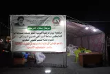 L'image montre une tente avec une grande banderole affichée en arabe et en français. La banderole aborde un message lié à un centre d'assistance. On peut voir des sacs et des fournitures au sol, suggérant que l'endroit est utilisé pour la collecte de dons ou l'aide humanitaire. L'éclairage est principalement nocturne, créant une ambiance sombre mais focalisée sur la tente.