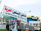 L'immagine mostra un grande striscione affisso su un edificio e altre insegne commerciali. Il striscione sembra contenere un messaggio che riguarda la libertà, con frasi in arabo e una richiesta di giustizia. Sullo sfondo, si intravedono alcuni edifici e attività commerciali, a suggerire un contesto urbano. L'ambiente appare vivace ma con un'atmosfera di forte espressione sociale.
