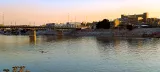 L'immagine mostra un paesaggio urbano al tramonto, con un fiume che riflette le sfumature del cielo. Sulla riva opposta del fiume ci sono edifici, alcuni dei quali sembrano avere una struttura moderna. In primo piano, un uomo sta remando su una piccola imbarcazione. Una passerella, o un ponte, si estende sopra il fiume, con lampioni che illuminano l'area. La scena trasmette un'atmosfera tranquilla, con colori caldi del cielo che sfumano verso l'orizzonte.