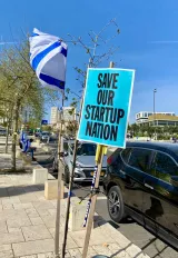 L'image montre un panneau à la couleur vive, turquoise, avec le texte "SAVE OUR STARTUP NATION" écrit en lettres blanches. À côté du panneau, on voit un drapeau, possiblement israélien, flottant dans le vent. L'arrière-plan présente une scène urbaine avec des voitures garées et des arbres. Le ciel est bleu, suggérant une journée ensoleillée. Cette image semble illustrer un rassemblement ou une manifestation, probablement en lien avec des enjeux économiques ou des préoccupations pour l'innovation dans le pays.