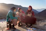 L'image montre un groupe de femmes assises ensemble dans un paysage montagneux. Elles semblent discuter tout en tenant des enfants dans leurs bras. La lumière du soleil brille, créant une atmosphère chaleureuse. Les femmes portent des vêtements traditionnels colorés, et le décor est naturel, avec des collines en arrière-plan. L'ambiance semble intime et paisible, suggérant un moment de partage et de connexion entre elles.