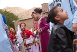 L'image montre un groupe de jeunes filles, vraisemblablement lors d'une fête ou d'un événement culturel. Elles portent des vêtements traditionnels colorés, avec des broderies vives. On peut voir des collines en arrière-plan, suggérant un cadre rural ou montagneux. Certaines filles semblent interagir entre elles, tandis que d'autres regardent autour, intriguées par leur environnement. L'atmosphère est festive et vivante.