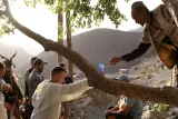 Sur l'image, on voit un groupe de personnes rassemblées dans un paysage rural montagneux. Un homme est assis sous un arbre, tandis qu'un autre, appuyé sur une branche, semble interagir avec lui ou passer quelque chose. L'atmosphère paraît conviviale, avec des montagnes en arrière-plan et une lumière douce qui illumine la scène. Les personnes sont habillées de façon traditionnelle, ce qui indique un contexte culturel spécifique.