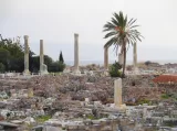 L'image montre des ruines archéologiques, avec plusieurs colonnes anciennes se dressant parmi un paysage de pierres et de décombres. Au premier plan, on peut voir des structures en pierre, tandis qu'à l'arrière-plan, quelques palmiers et arbres ajoutent une touche de verdure. Le ciel est légèrement nuageux, et l'horizon laisse entrevoir la mer. L'ensemble évoque un site historique riche en culture et en histoire.