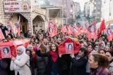 L'image montre une grande foule rassemblée devant un bâtiment, possiblement un centre culturel ou éducatif. Les participants tiennent des drapeaux rouges ornés de portraits, tandis que certains portent une grande caisse ou un cercueil, symbolisant probablement une commémoration. Les visages des personnes expriment des émotions fortes, telles que la tristesse ou la colère. L'atmosphère générale semble être celle d'un rassemblement politique ou d'une manifestation pour honorer quelqu'un.