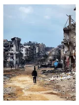 L'image montre un paysage de destruction. Au centre, une route en terre mène à travers des bâtiments effondrés et des débris, témoignant d'une grande dévastation. Quelques personnes marchent au loin, semblant solitaires dans cet environnement désolé. Le ciel est nuageux, ajoutant une atmosphère sombre à la scène. Les ruines des structures et le manque de végétation évoquent un lieu de conflit ou de catastrophe.