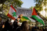 L'image montre une manifestation avec des drapeaux du Liban et de la Palestine. Au premier plan, une personne tient une pancarte sur laquelle est écrit "KHALAS". En arrière-plan, on peut voir d'autres manifestants et un environnement urbain avec des arbres. L'atmosphère semble engagée et solidaire.