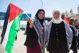 L'image montre deux femmes représentant une scène de manifestation ou de rassemblement. Elles portent des vêtements traditionnels et des foulards. L'une d'elles tient un drapeau palestinien, qui est tricolore avec du noir, du rouge, du vert et du blanc. L'arrière-plan laisse entrevoir une rue avec quelques personnes et des enseignes. L'atmosphère semble être festive et engagée.