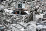 L'image montre des débris d'un bâtiment effondré, créant un paysage chaotique. Au milieu des ruines, on aperçoit une voiture, à l'intérieur de laquelle se trouvent deux personnes. La voiture semble avoir été prise au piège dans les décombres, et l'environnement environnant est marqué par la destruction. Des morceaux de béton et d'autres matériaux de construction sont éparpillés autour de la scène, accentuant l'impact d'une catastrophe.