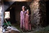 L'image montre deux jeunes filles se tenant côte à côte dans un intérieur rustique. Elles portent des robes simples aux teintes douces, et leur expression semble contemplative. L'ambiance de la scène est sombre, avec des murs en pierre et des éléments naturels tels que de l'herbe au sol. Une lumière douce pénètre par une fenêtre, créant un contraste avec l'ombre de la pièce. L'ensemble dégage une atmosphère de simplicité et d'authenticité.