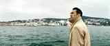 L'image montre un homme debout près d'une étendue d'eau, probablement une mer ou un lac. Il regarde au loin, vers une ville côtière qui s'étend sur l'horizon, avec des bâtiments colorés nichés sur les collines. Le ciel est nuageux, ce qui donne une ambiance un peu mélancolique à la scène. L'homme porte un trench-coat beige, ce qui suggère qu'il fait frais. L'environnement naturel et urbain se mélange harmonieusement, créant une atmosphère contemplative.