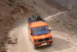 L'image montre un véhicule orange, probablement un fourgon, circulant sur un chemin de terre étroit et sinueux en montagne. À l'arrière du véhicule, plusieurs personnes sont assises, probablement en train de profiter de la vue. Le paysage environnant est sec, avec des rochers et quelques collines. Le ciel semble dégagé, ce qui suggère une belle journée pour voyager.