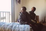 Deux hommes assis sur un lit, l'un souriant, l'autre regardant par la fenêtre.