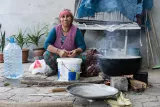 L'image montre une femme assise à l'extérieur, au milieu d'un espace de travail rustique. Elle porte un foulard sur la tête et un gilet coloré. Devant elle, il y a une grande marmite sur un feu, émettant de la vapeur. À côté, on aperçoit un seau et une bouteille d'eau. Le fond est décoré de quelques plantes en pots et d'un abri en toile. L'ensemble de la scène évoque une ambiance traditionnelle et artisanale.