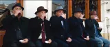 L'image montre un groupe de cinq hommes assis côte à côte, tous portant des chapeaux. Ils semblent apprécier une boisson, probablement du thé, en levant leurs tasses. Ils sont vêtus de manteaux sombres et semblent être dans un environnement extérieur, probablement sur une terrasse ou devant un café. L'atmosphère dégage une sensation de convivialité et de camaraderie.
