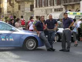 L'image montre plusieurs policiers en uniforme se tenant près d'une voiture de police. Ils semblent discuter ou se détendre dans une rue animée, entourés de passants qui se déplacent. Les bâtiments en arrière-plan sont typiques d'une ville européenne, avec des façades anciennes et des boutiques. L'atmosphère semble vivante et dynamique, caractéristique d'une zone touristique.