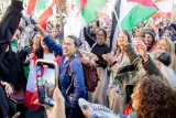 L'image montre une foule de personnes rassemblées, manifestant avec des drapeaux de différents pays, notamment le Liban et l'Algérie. Les participants semblent enthousiastes et engagés, certains dansent et chantent, tandis que d'autres applaudissent. L'ambiance est festive et dynamique, reflétant un esprit de solidarité et de célébration.