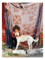L'image montre un jeune enfant avec des cheveux bouclés, qui se tient près d'un chien de couleur blanche. L'enfant semble heureux et souriant, enlaçant le chien. En arrière-plan, il y a un mur fait de tapis ou de tissus multicolores qui servent de toile de fond. L'ambiance générale est chaleureuse et affectueuse, mettant en valeur le lien entre l'enfant et le chien.