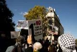 L'image montre une manifestation dans une rue animée. Au premier plan, une personne tient une pancarte qui indique "1 + 80 ANS DE GÉNOCIDE / AN DE RÉSISTANCE". En arrière-plan, on aperçoit d'autres manifestants portant des slogans et des affiches en rapport avec des questions sociales et politiques, tandis qu'un bâtiment caractéristique se dresse à côté. Le ciel est dégagé et la lumière du jour met en valeur l'atmosphère dynamique de l'événement.