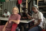 L'image montre un groupe de personnes dans un intérieur, probablement dans un centre communautaire ou une salle de classe. Une femme au premier plan, portant des lunettes et une blouse à pois, sourit à la caméra. À l'arrière, deux jeunes hommes sont assis autour d'une table. L'un semble regarder son téléphone, tandis que l'autre fait un signe de la main. Un troisième homme est également présent, apparu au fond de la pièce. L'ambiance semble détendue et conviviale.
