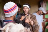 L'image montre un groupe d'hommes en train d'interagir dans un espace intérieur. Certains d'entre eux portent des vêtements faits de fourrure ou de poils, ce qui évoque une tradition culturelle ou une pratique artisanale. L'un des hommes est en cours d'habillage ou d'ajustement d'un vêtement, tandis que les autres observent. L'environnement est rustique, avec des murs en bois et des détails simples. L'atmosphère semble conviviale et centrée sur le partage d'une activité sociale.