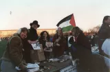 L'image montre un groupe de personnes lors d'un événement extérieur, probablement une manifestation ou une rassemblement. À l'avant-plan, plusieurs personnes souriantes se tiennent derrière une table couverte de brochures et de pamphlets. L'une des femmes tient un drapeau palestinien, ce qui suggère un soutien à une cause. L'arrière-plan montre une foule et un paysage urbain, avec des arbres et des bâtiments. L'atmosphère semble amicale et engagée, semblant refléter une mobilisation pour une cause sociale ou politique.