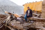 L'image montre un homme assis sur les débris d'une structure, probablement après un événement destructeur. Il est vêtu d'une veste sombre et semble pensif. En arrière-plan, on aperçoit des montagnes et des éléments de la nature, tandis qu'un tissu jaune pend au-dessus de lui, ajoutant une touche de couleur à l'environnement. Le site apparaît en désordre, avec des matériaux éparpillés, suggérant des dégâts. L'ensemble dégage une atmosphère de résilience face à l'adversité.