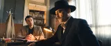 L'image montre deux hommes assis à une table dans un bureau. L'un d'eux est vêtu d'un costume noir et porte un chapeau, semblant sérieux et pensif. L'autre homme, en tenue plus décontractée, semble l'observer attentivement, peut-être en train de poser des questions. La lumière d'une lampe éclaire la scène, créant une atmosphère tendue et intrigante. Les murs sont décorés de manière simple, et on peut apercevoir des dossiers ou des documents en arrière-plan, suggérant un cadre professionnel.