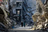 L'image montre une rue dévastée, remplie de débris et de bâtiments effondrés. Au premier plan, deux femmes marchent côte à côte. L'atmosphère est sombre et désolante, illustrant les conséquences visibles d'une destruction massive. Les murs des bâtiments sont endommagés, et des morceaux de béton jonchent le sol.