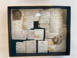 L'image montre une boîte encadrée contenant plusieurs morceaux de papier ou cartes de différentes tailles et formes. Ces papiers sont couverts de notes, de ratures et d'écritures dans plusieurs langues, y compris des caractères arabes et latins. On peut également apercevoir une vieille photographie en noir et blanc d'un homme. L'ensemble présente un aspect ancien et nostalgique, avec des traces de temps visibles sur les papiers. La disposition des éléments semble aléatoire, créant un collage d'histoires et de souvenirs.