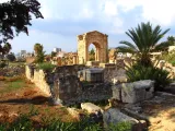 L'image montre des ruines antiques, probablement d'un site archéologique. On peut voir de grands murs en pierre, des colonnes et des structures partiellement effondrées, entourées de végétation comme des palmiers et des arbustes. L'arrière-plan présente un ciel nuageux, et on aperçoit des bâtiments modernes au loin, ce qui contraste avec l'ancienneté des ruines. L'ensemble évoque un lieu chargé d'histoire, invitant à la contemplation.