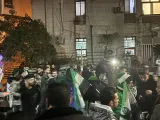 L'image montre une foule de personnes rassemblées dans une rue, portant des drapeaux syriens. Il y a des bâtiments en arrière-plan et une ambiance festive, avec des lumières et des visages souriants. Des gens semblent échanger et célébrer ensemble.