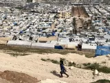 L'image montre un paysage vaste où l'on peut voir de nombreuses habitations regroupées, probablement des abris ou des tentes, disposées de manière ordonnée. Au premier plan, un enfant court sur un terrain montagneux, accentuant le contraste entre son innocence et la dureté de l'environnement autour de lui. En arrière-plan, on aperçoit une grande étendue de toits de tentes, indiquant une zone de peuplement dense, ce qui suggère un contexte de crise ou de déplacement. La scène évoque des sentiments de résilience et de survie dans des conditions difficiles.