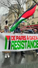 L'image montre un groupe de manifestants lors d'une marche, probablement en faveur de la Palestine. Ils portent une grande bannière exprimant le message "De Paris à Gaza" et "Résistance", avec des couleurs vives. Des drapeaux palestiniens et peut-être un drapeau noir, rouge et jaune sont également visibles. Les participants semblent déterminés et solidaires, malgré le temps pluvieux suggéré par les vêtements et l'environnement.