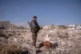 L'image montre un homme en uniforme militaire se tenant debout dans un paysage de ruines, avec des bâtiments détruits en arrière-plan. Le sol est rempli de débris, et l'environnement est aride et désertique. L'homme porte un fusil et regarde vers l'horizon.
