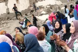L'image montre un groupe de femmes rassemblées, chantant et dansant joyeusement. Certaines tiennent des tambours ou des instruments de musique, tandis que d'autres applaudissent. Les expressions sur leurs visages montrent de la joie et de l'énergie. À l'arrière-plan, on peut voir des enfants et des adultes se mêlant à l'événement, avec des ruines en pierre. L'atmosphère semble festive et communautaire, reflétant une célébration culturelle ou une réunion sociale. La diversité des vêtements et des couleurs ajoute à la richesse de la scène.