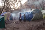L'image montre un campement composé de plusieurs tentes colorées, disposées de manière relativement désordonnée. On peut voir des arbres dénudés en arrière-plan, et la lumière du soleil perce à travers les branches, créant une atmosphère mélancolique. Des enfants sont présents dans la scène, dont certains semblent s'occuper de tâches quotidiennes. De la fumée s'échappe d'un feu improvisé, suggérant que les habitants préparent de la nourriture ou se réchauffent. L'environnement semble rural et un peu désolé, témoignant d'une situation de précarité.