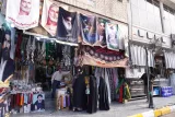 L'image montre une rue animée remplie de boutiques. À l'avant-plan, on peut voir un magasin décoré d'affiches, probablement de personnalités religieuses ou historiques, et des articles tels que des vêtements et des accessoires. Des personnes sont visibles, certaines portant des vêtements traditionnels. À l'arrière-plan, on aperçoit d'autres bâtiments. L'atmosphère semble vivante et culturelle.