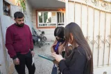 L'image montre trois personnes dans un environnement extérieur, probablement devant une maison. Un homme est debout à gauche, vêtu d'un pull bordeaux. Il semble observer attentivement les deux femmes qui se trouvent à sa droite. Ces dernières portent des masques et consultent quelque chose sur une tablette. L'arrière-plan montre un espace de jardin et un vélo. L'atmosphère paraît concentrée, peut-être liée à une discussion ou à une collecte d'informations.