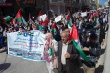 L'image montre une manifestation avec un grand groupe de personnes marchant dans la rue, brandissant des drapeaux palestiniens. Au premier plan, on voit des individus portant des pancartes et des banderoles avec des messages en arabe. L'atmosphère semble être celle d'une mobilisation pacifique avec des participants vêtus de manière variée. Des journalistes filment l'événement. Les bâtiments en arrière-plan et la disposition des personnes suggèrent une ambiance dynamique et engagée.