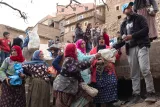 L'image montre un groupe de femmes, probablement dans un village, rassemblées autour d'un homme qui semble distribuer quelque chose. Les femmes portent des vêtements traditionnels et des foulards colorés, et certaines semblent engager une conversation animée. En arrière-plan, on aperçoit des bâtiments en terre et des habitants qui observent la scène, créant une atmosphère communautaire. Les expressions des femmes varient entre curiosité et enthousiasme. Cela évoque un moment de partage et de solidarité dans leur communauté.