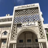 L'image montre un bâtiment architectural imposant qui semble être un édifice public. La façade est ornée de motifs géométriques qui ajoutent une touche esthétique. Au-dessus de l'entrée, on peut distinguer des inscriptions en arabe. Le bâtiment dégage une impression d'ancienneté, avec des éléments architecturaux traditionnels et une structure en pierre. Le ciel est bleu, laissant entrevoir une belle journée.