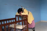 L'image montre une femme habillée d'un manteau jaune et d'une jupe rose, exprimant une attitude de fatigue ou de tristesse. Elle est penchée sur un lit à barreaux en bois, avec un coussin dessus. La pièce est peinte en bleu, mais on peut voir des fissures sur le mur. L'atmosphère semble réfléchissante et émotive, suggérant un moment de contemplation ou de désespoir.