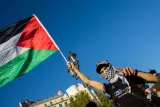 L'image montre un homme portant un T-shirt avec un logo, brandissant fièrement un drapeau palestinien. Il porte également un foulard à motif checkered qui couvre une partie de son visage. Le ciel est d'un bleu clair, et l'ambiance semble être celle d'une manifestation ou d'un rassemblement, avec des bâtiments en arrière-plan. L'homme fait un signe de paix avec sa main, ajoutant à l'esprit de solidarité de la scène.